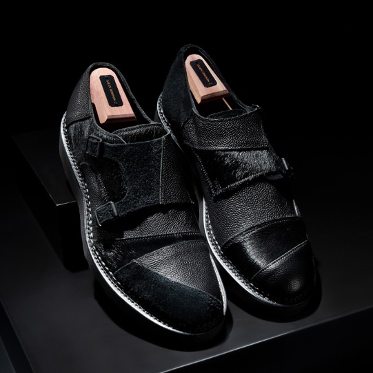 A pair of brown dress shoes with a burnished finish, shown on a dark background. One shoe is displayed upright and the other lying on its side to reveal the polished leather sole.