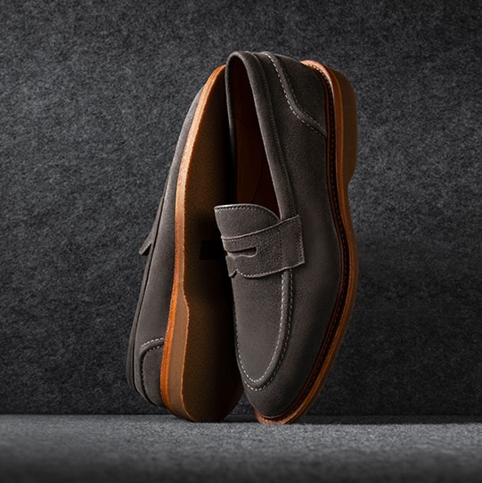 A pair of brown dress shoes with a burnished finish, shown on a dark background. One shoe is displayed upright and the other lying on its side to reveal the polished leather sole.