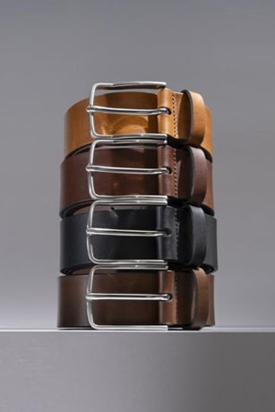 Four men’s leather belts stacked on top of one another with a gray background.  