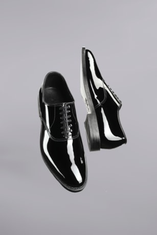 A pair of black men’s patent leather dress shoes floating in the air with a gray background. 