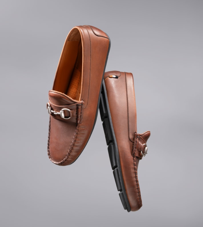 A pair of men’s brown leather bit loafers floating in the air with a gray background. 