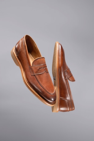 A pair of men’s brown penny loafers floating in the air with a gray background.  