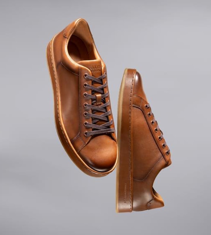 A pair of men’s brown leather sneakers floating in the air with a gray background. 