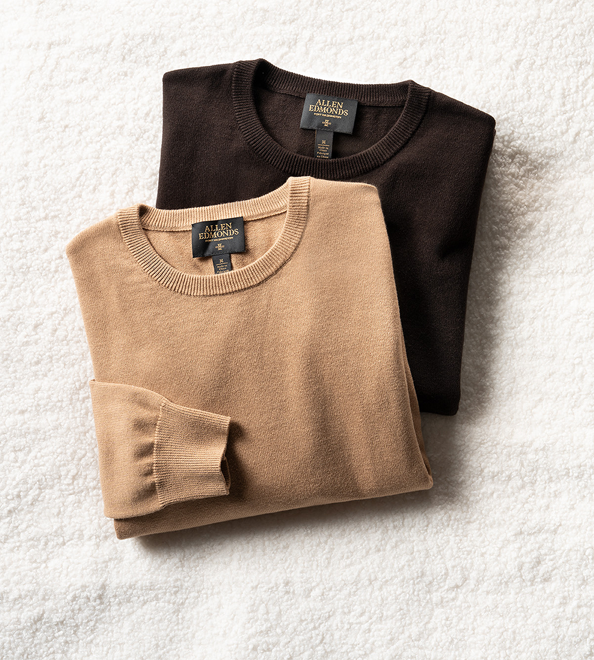 Two folded men’s sweaters overlapping each other, one in a tan color and one in dark brown