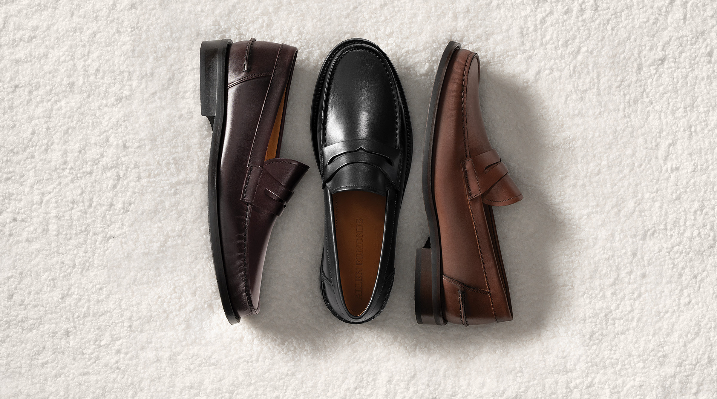 Three men’s penny loafers next to each other, one in black leather and two in brown leather. The brown loafers are laying on their side, while the black loafer is in the middle, laying upright.  