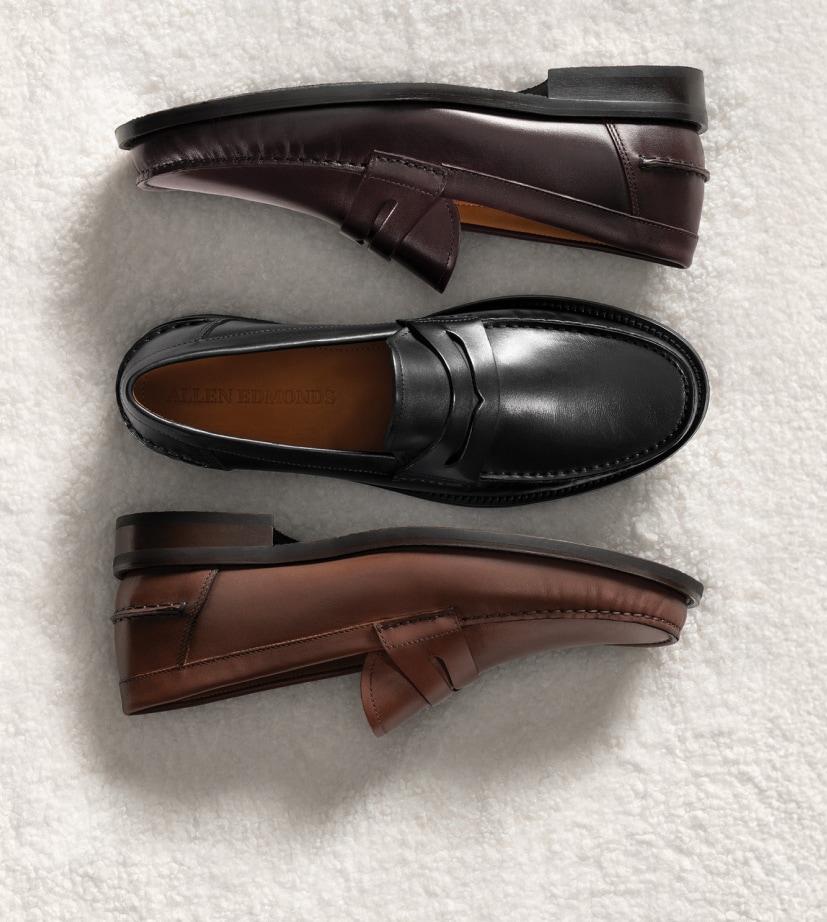 Three men’s penny loafers next to each other, one in black leather and two in brown leather. The brown loafers are laying on their side, while the black loafer is in the middle, laying upright.  