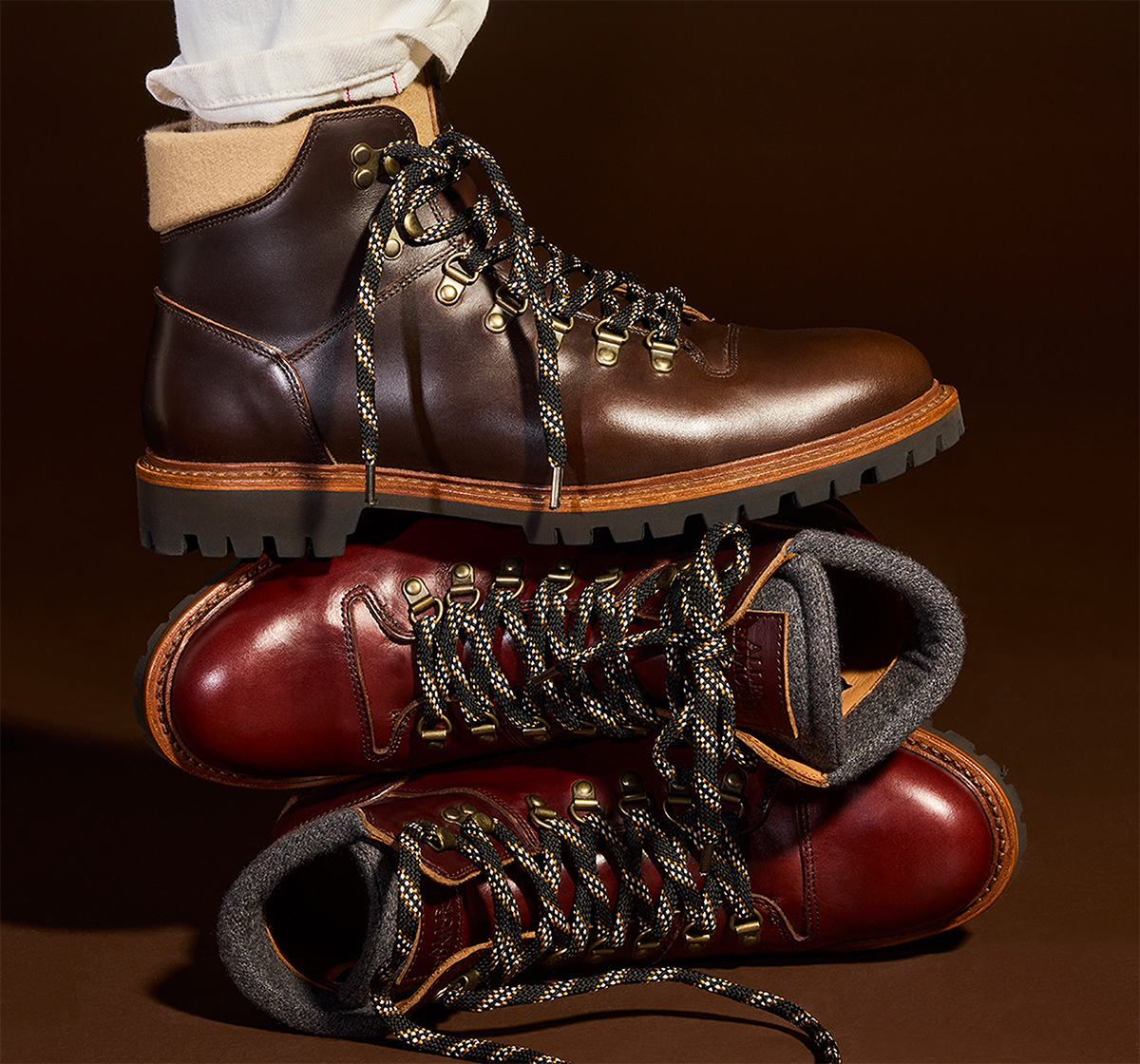 A man sitting down wearing a black shearling-lined leather jacket, gray turtleneck sweater, and white pants.