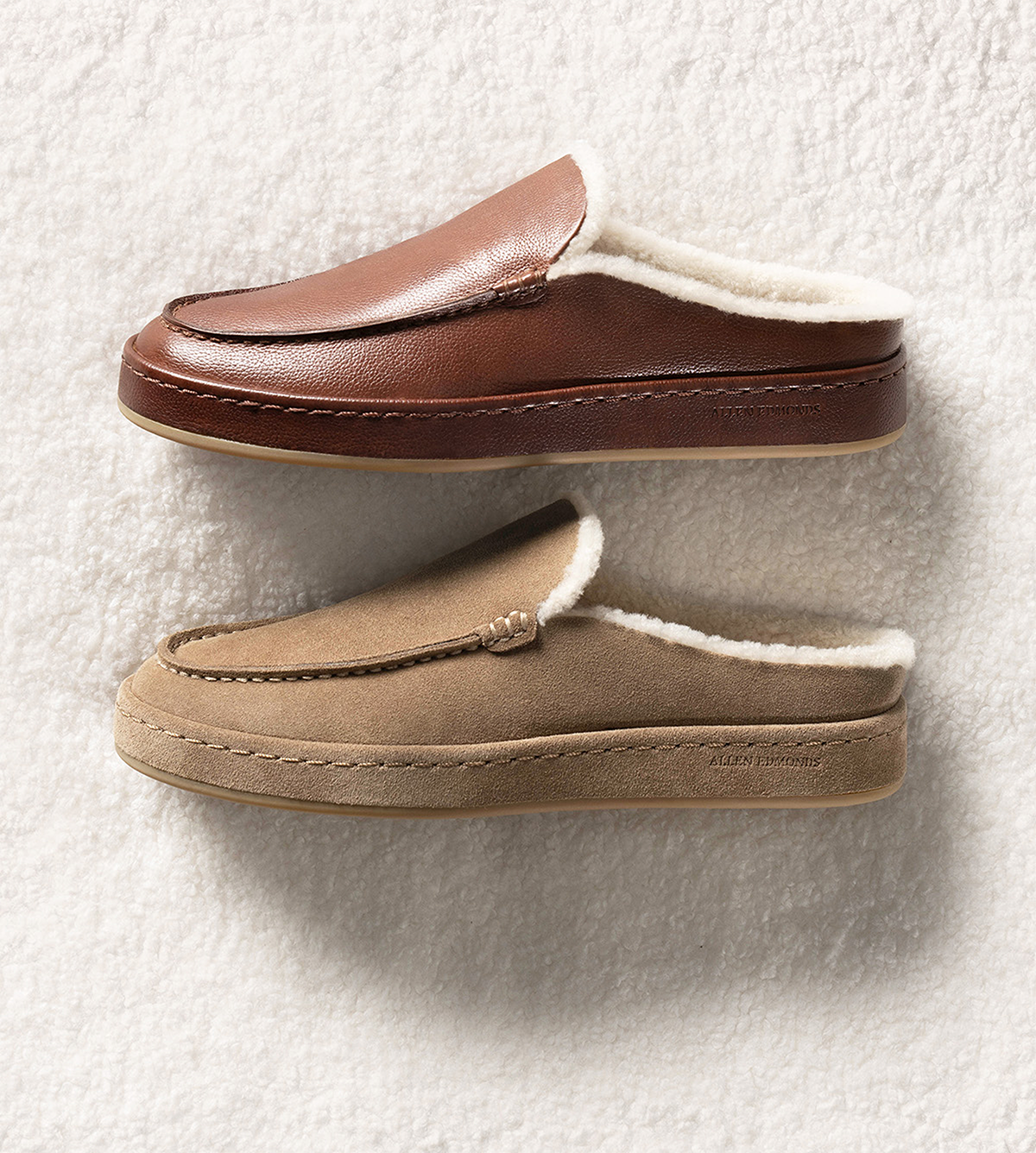 Two shearling-lined men’s slippers displayed laying on their side. One is in brown leather, and the other is tan suede. 