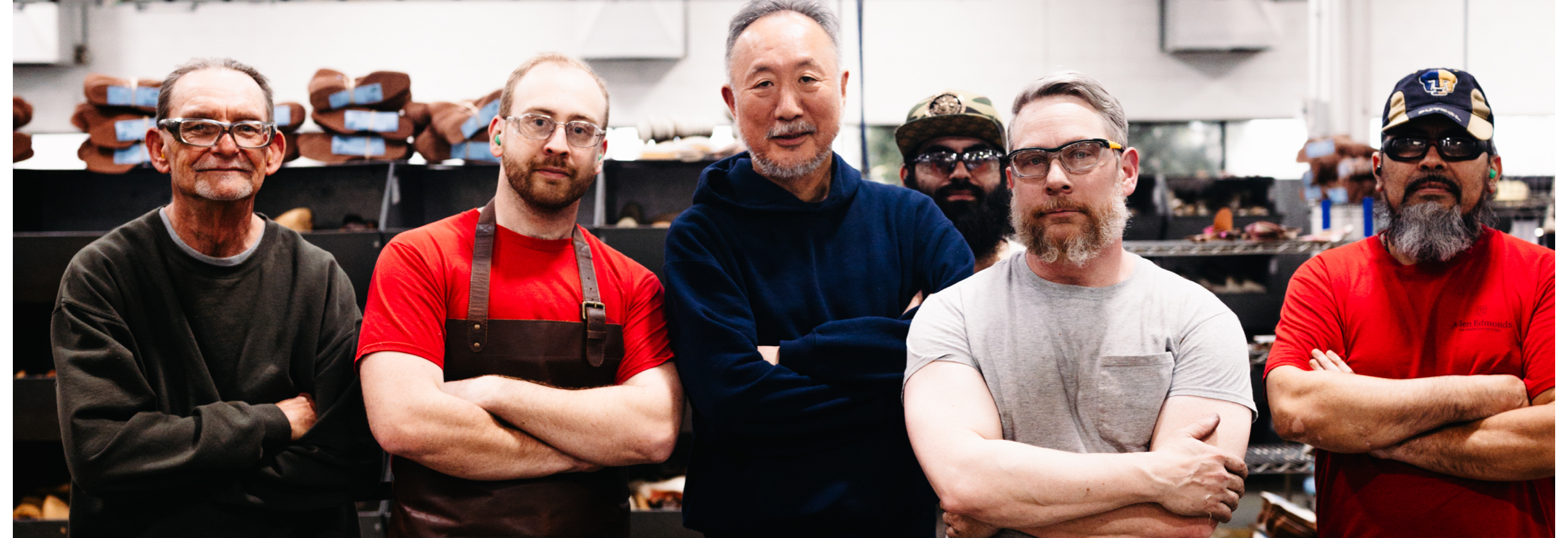 Daiki Suzuki and his team standing in a shoe workshop with arms crossed, with shelves of leather materials and tools in the background.