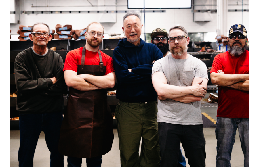 Daiki Suzuki and his team standing in a shoe workshop with arms crossed, with shelves of leather materials and tools in the background.