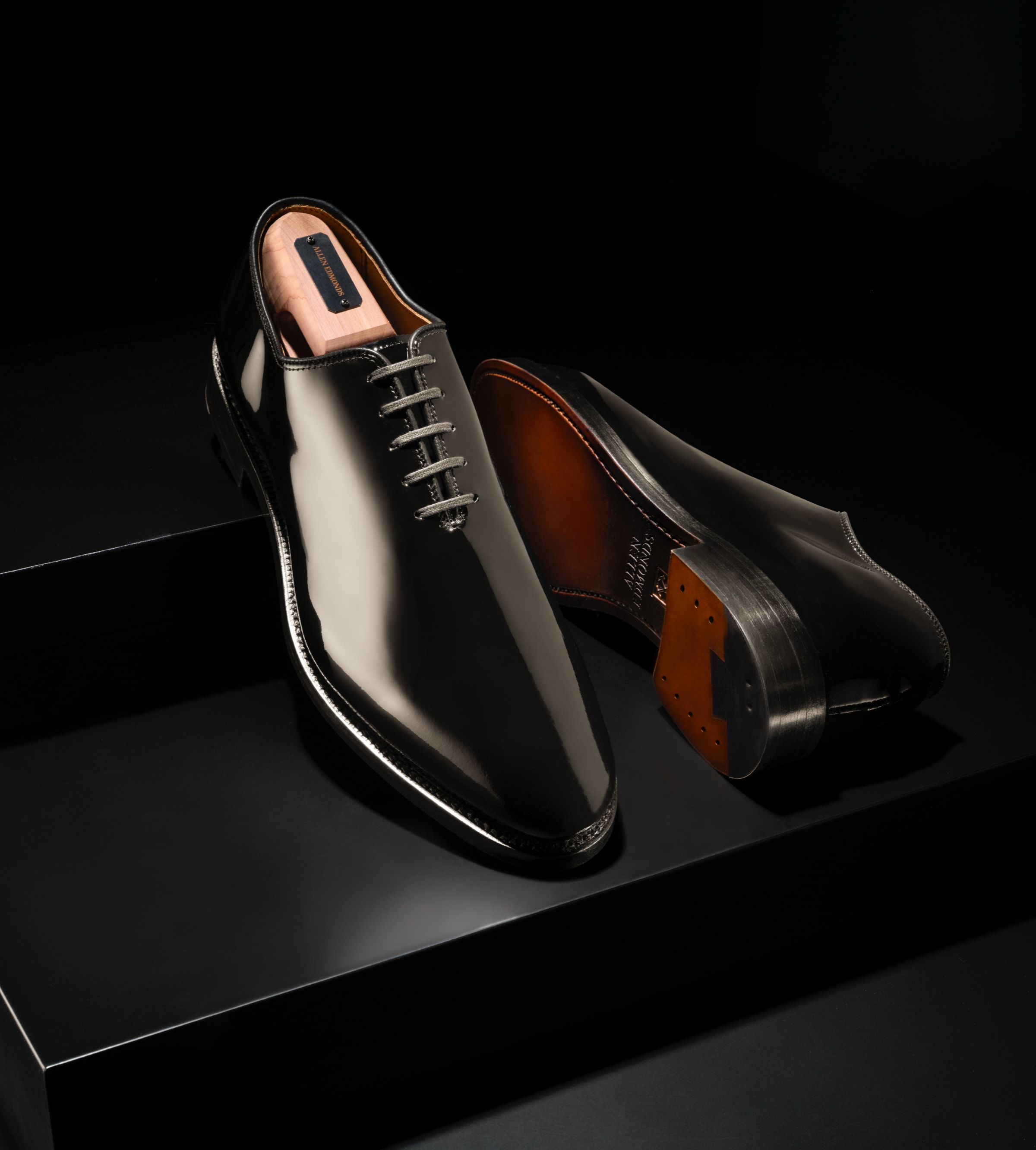A pair of men’s black patent leather Oxford dress shoes with one laying on its side. 