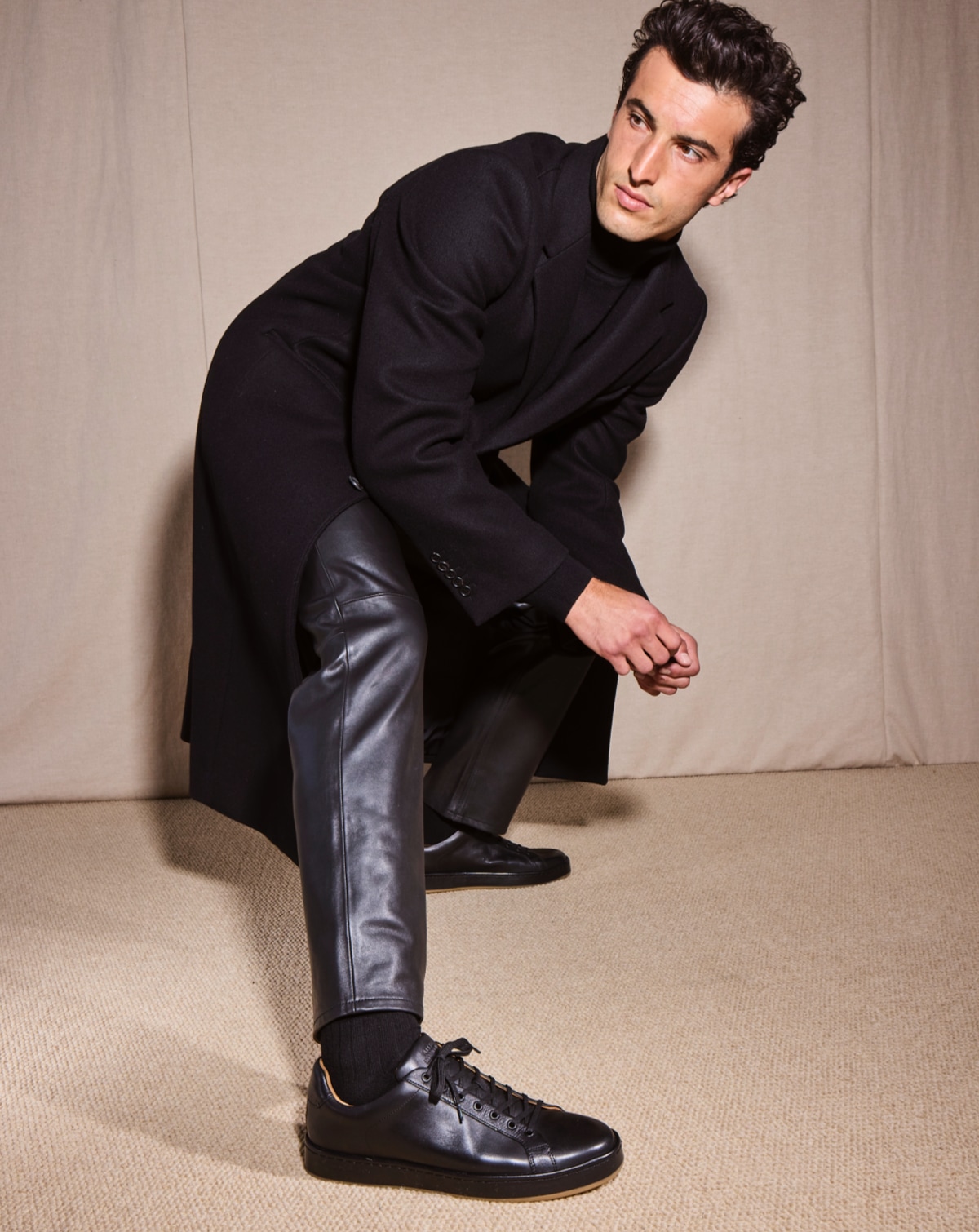 A male model wearing an all-black outfit featuring a pair of Allen Edmonds Barnes Lace Up Sneakers in black leather.  