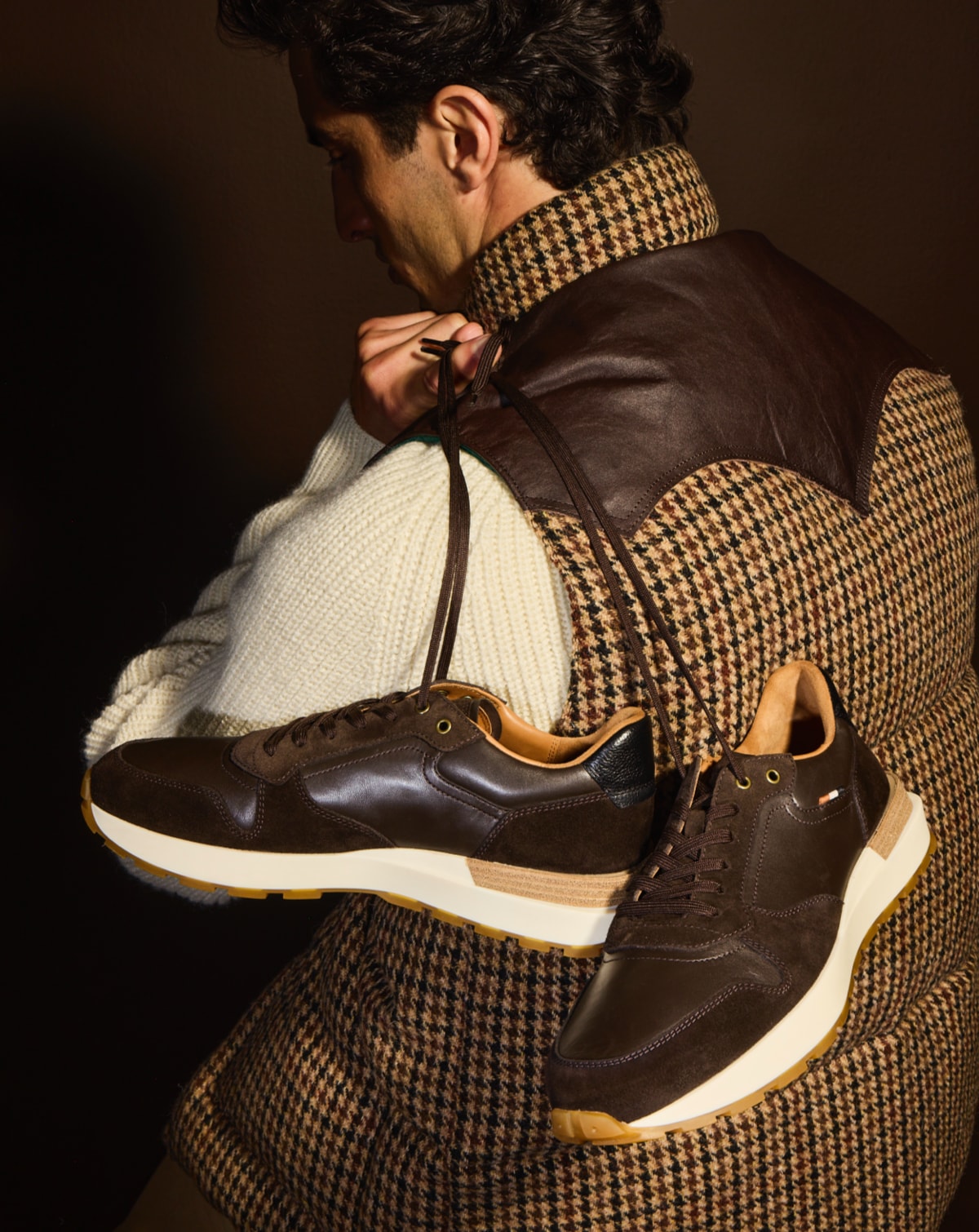 A male model holding a pair of brown suede lace up sneakers by the laces over his shoulder.