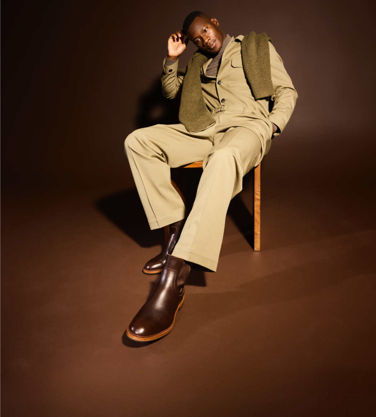 A man sitting in a chair modeling a tan pair of pants and matching jacket, with a green sweater draped over his shoulder and a pair of brown leather Chelsea boots.