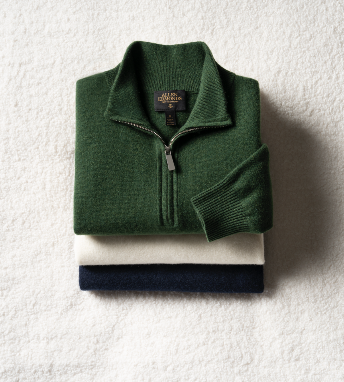 Three men’s zip up sweaters folded and stacked on top of one another. One is green, one is white, and one is navy blue.  