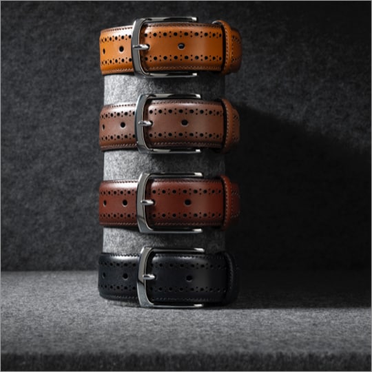 Four men’s leather belts stacked on top of one another. One is black and the other three are various shades of brown.   