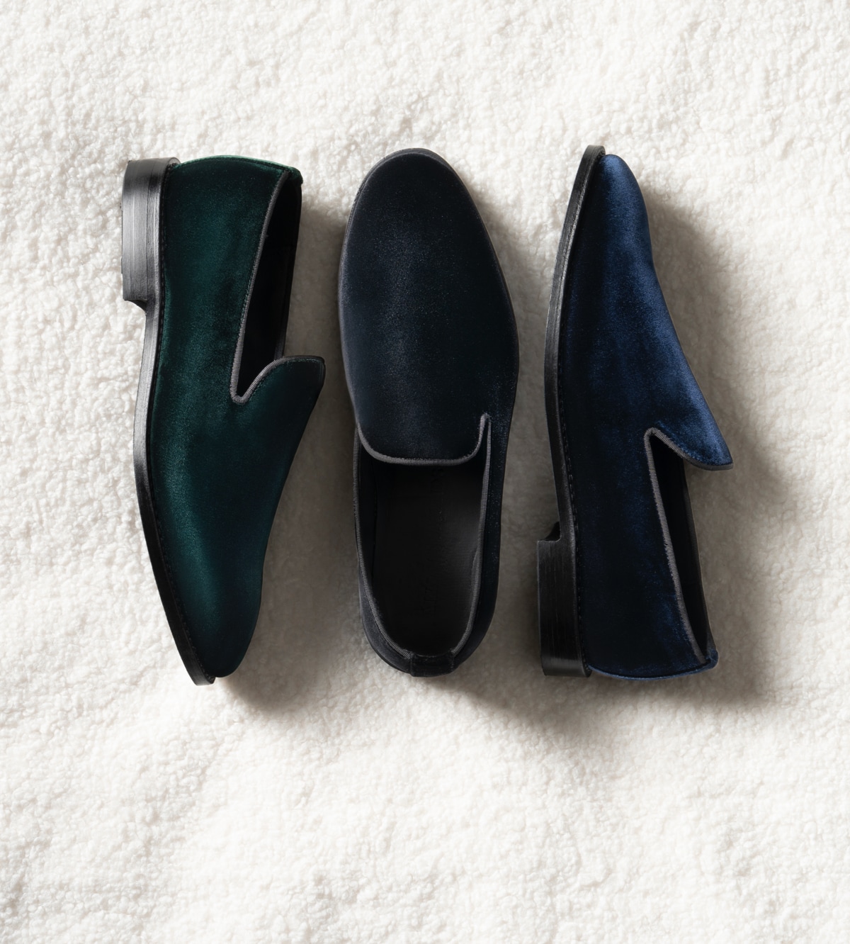 A top view of three velvet loafers. One is black, one is blue, and one is green. The black one is displayed on its sole, while the green and blue shoes are displayed on their side. 