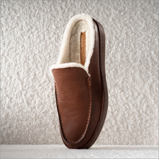 A men’s slipper in brown suede with white shearling lining displayed vertically on its toe.  