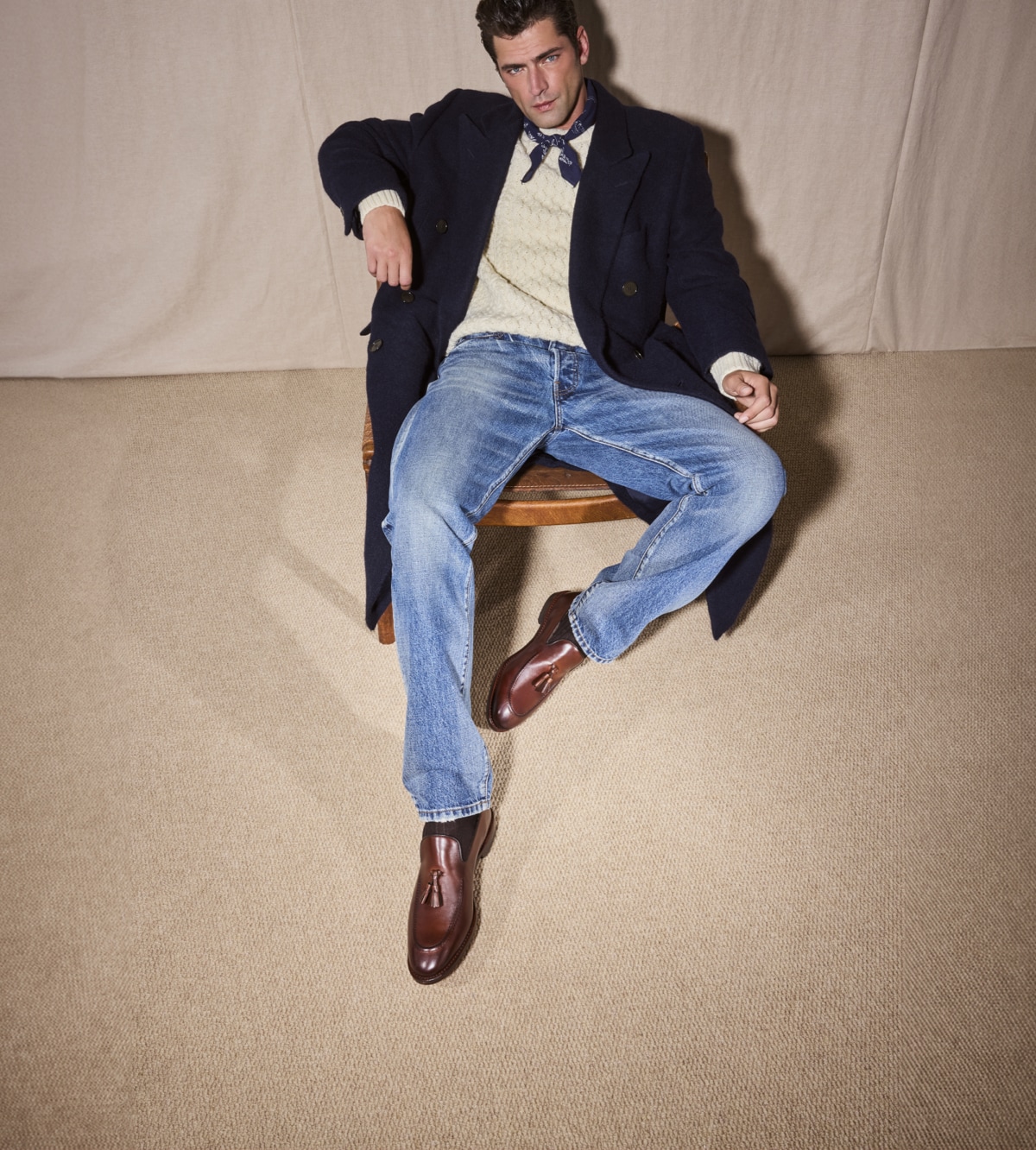 A man sitting in a chair wearing a black pea coat, an off-white sweater, jeans, and a pair of brown leather tassel loafers.