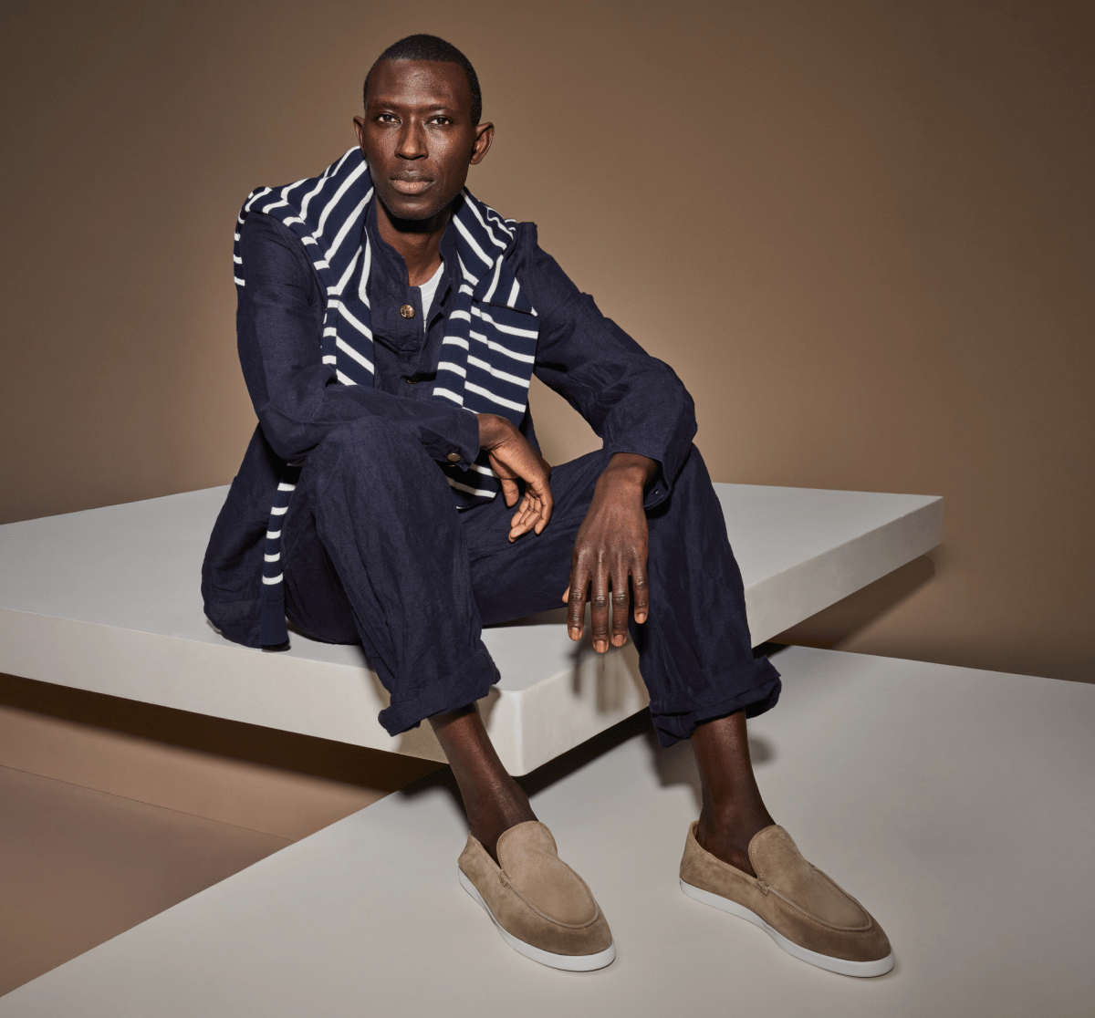 Man seated on white platform wearing Taupe Suede Gino Loafers, styled with a navy linen suit and navy and white striped scarf. 