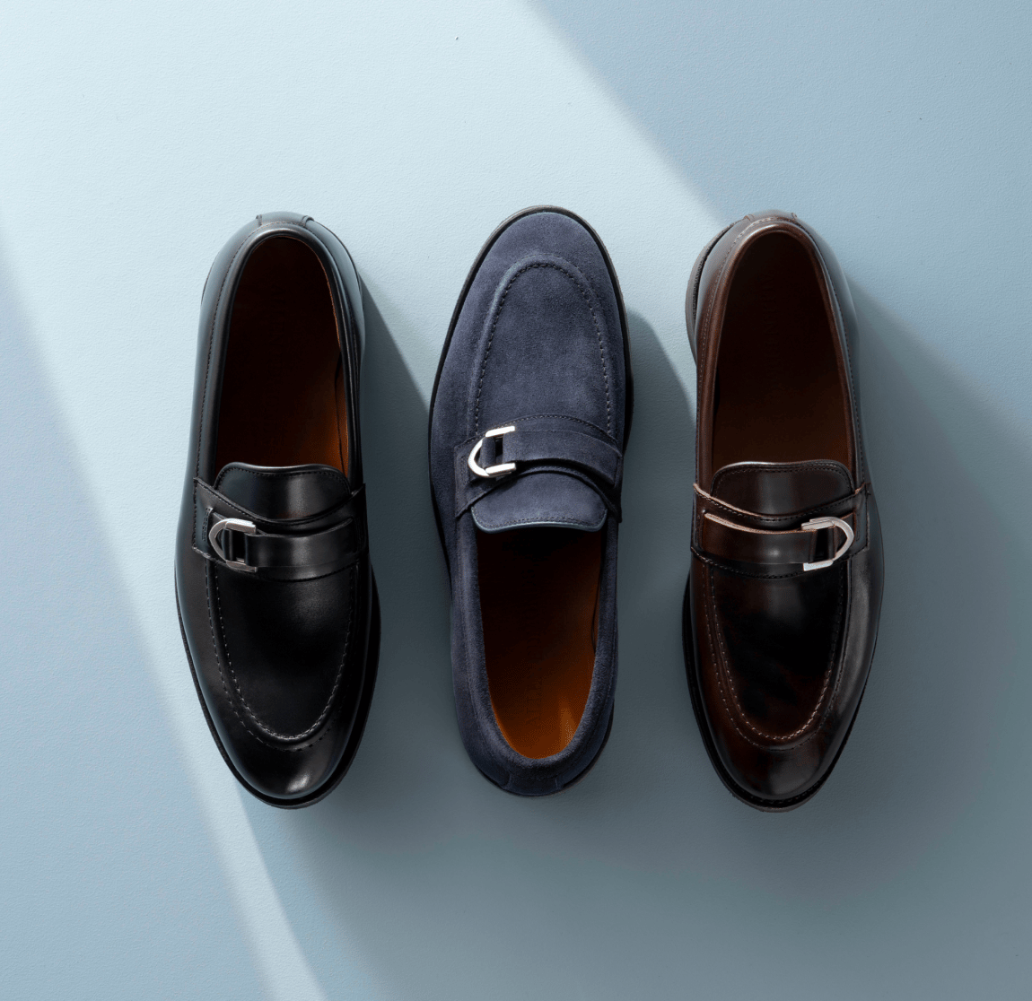 Dark brown, black, and navy leather Allen Edmonds Rollins buckle loafers with silver buckles against a light blue background. 