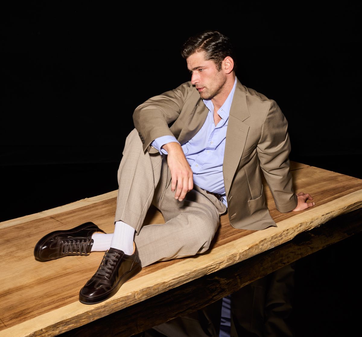 Dark brown Allen Edmonds Hawthorne sneakers and white socks worn by a man in a tan suit sitting on a wooden platform 