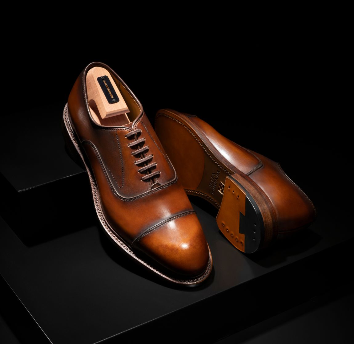 Brown leather Allen Edmonds Manhattan Cap-toe oxford dress shoes displayed with a branded cedar shoe tree against a dark background. 