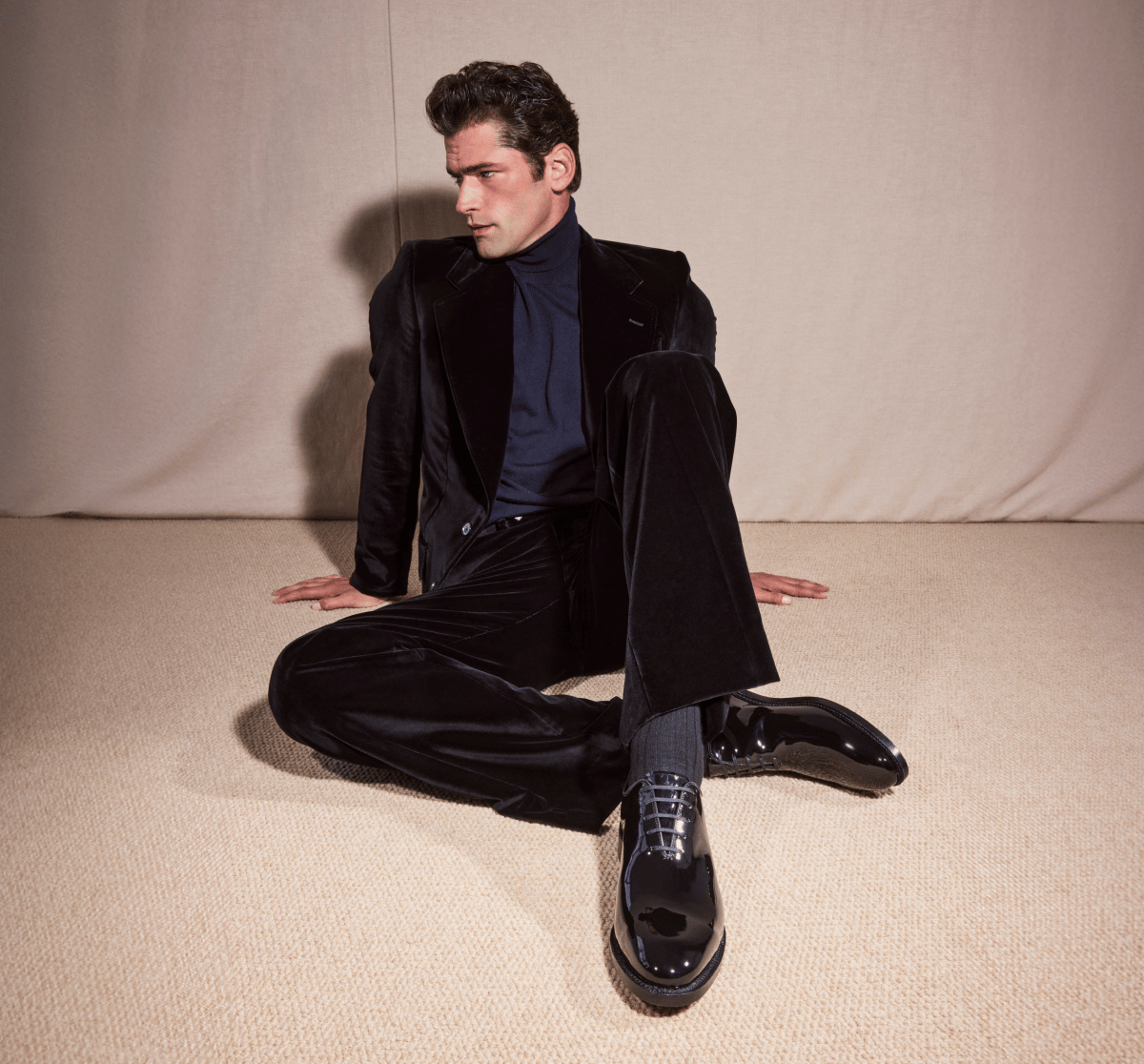 Black patent leather Allen Edmonds oxford dress shoes and dark socks worn by a man in a black velvet suit and navy turtleneck sitting on a neutral carpet 