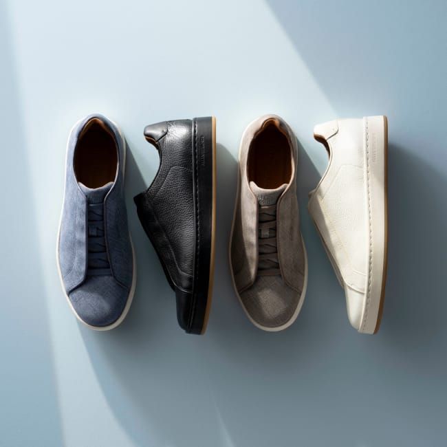 Top-down view of blue, black, grey, and white Allen Edmonds Banks stretch-lace sneakers with tan linings against a light blue background.