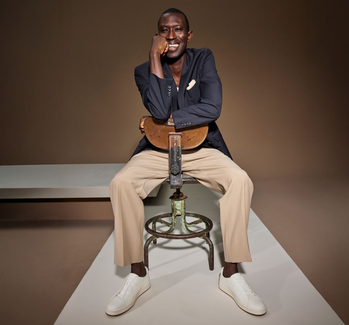 White leather Allen Edmonds Banks stretch-lace sneakers worn by a man in a navy pinstripe blazer and tan trousers sitting on a vintage metal stool