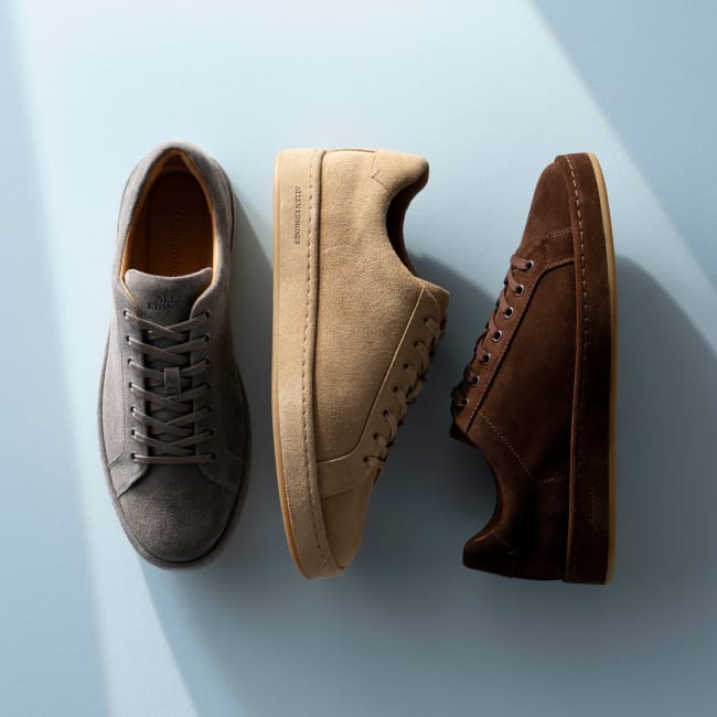 Grey, tan, and dark brown suede Allen Edmonds Barnes sneakers with matching color laces against a light blue background. 