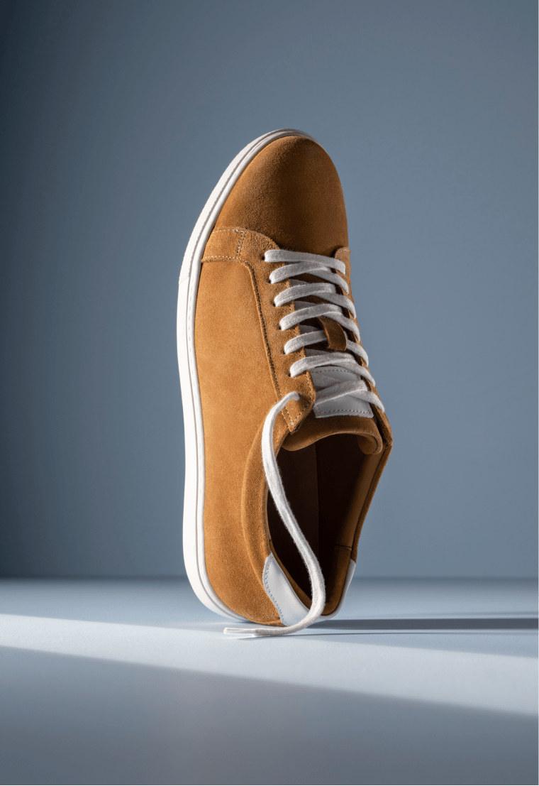 Tan suede Allen Edmonds Courtside sneaker with white laces and a white sole against a light blue background. 