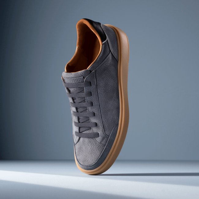 Grey leather Allen Edmonds Oliver stretch-lace sneaker with charcoal laces, tan lining, and a gum sole against a light blue background. 