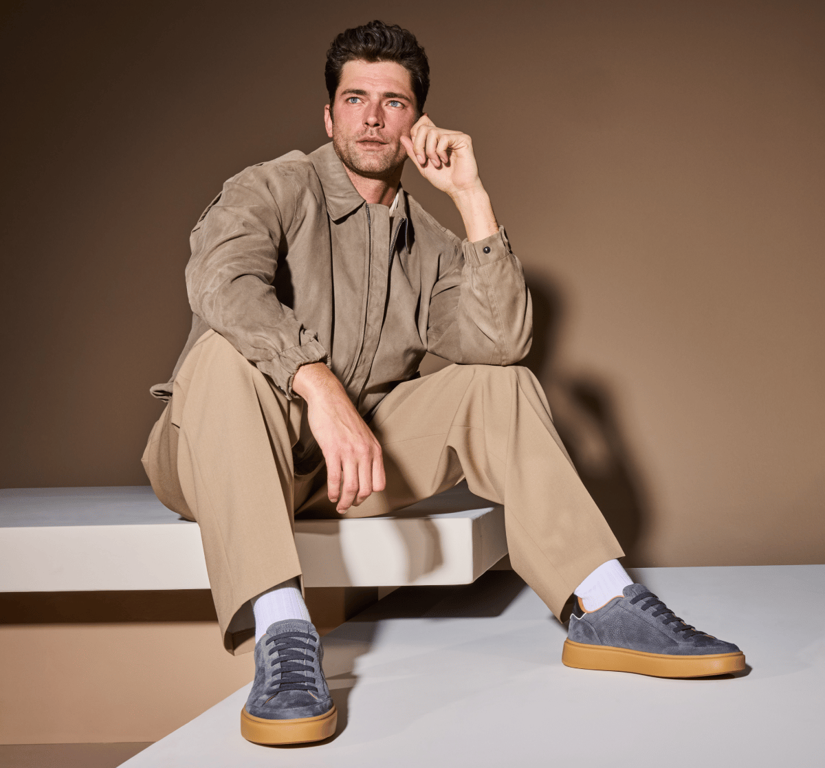 Grey suede Allen Edmonds Oliver stretch-lace sneakers with charcoal laces worn by a man in a tan bomber jacket and khaki trousers sitting on a white platform.