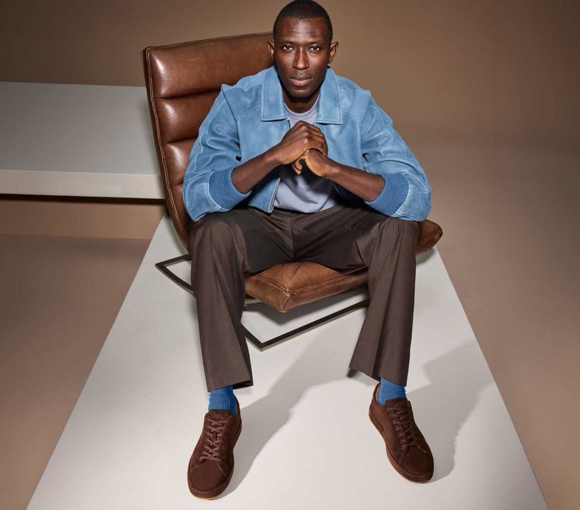 A man sitting in a leather chair wearing the brown suede Barnes Lace-up sneaker with brown pants and a blue jacket.