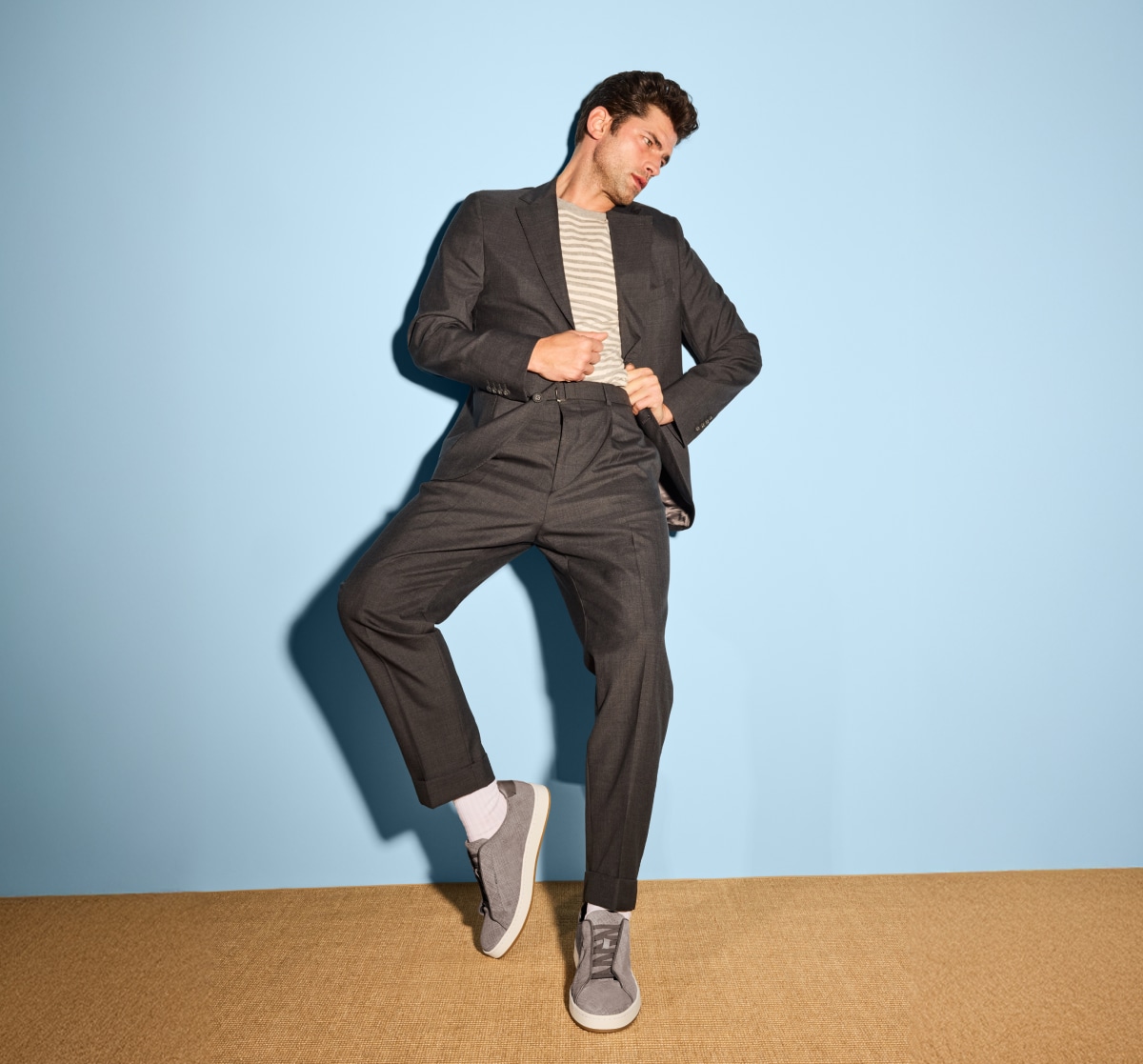 Man wearing Allen Edmonds Navy Suede Banks Stretch-Lace Sneakers, styled with a charcoal suit and striped crewneck sweater against a light blue background 